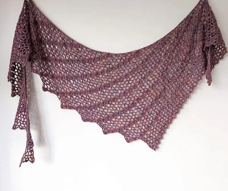 An asymmetrical mauve crochet shawl featuring a delicate, open lace pattern with repeating V-stitches and shell rows. The shawl has a scalloped bottom edge and is made from fine fingering-weight yarn.