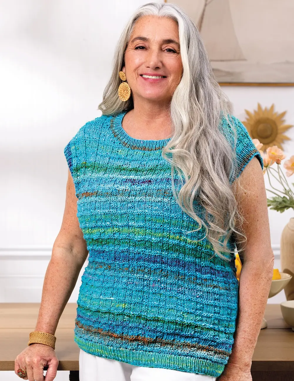 A woman with long, wavy grey hair is smiling at the camera. She is wearing a teal and blue sleeveless, knitted vest with horizontal stripes.