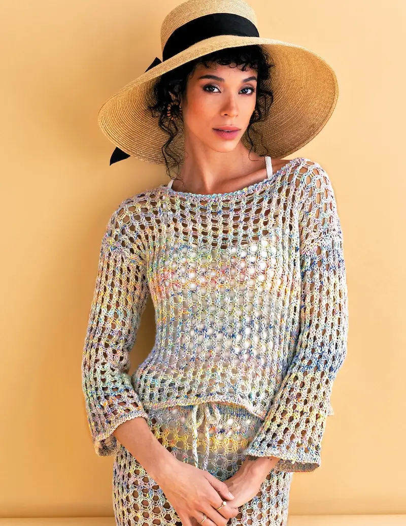 A woman with dark, loosely pulled-back hair models a matching two-piece set, consisting of a short-sleeved top with a boat neck and a knee-length skirt. Both pieces feature an open, multi-colored mesh knit design. 