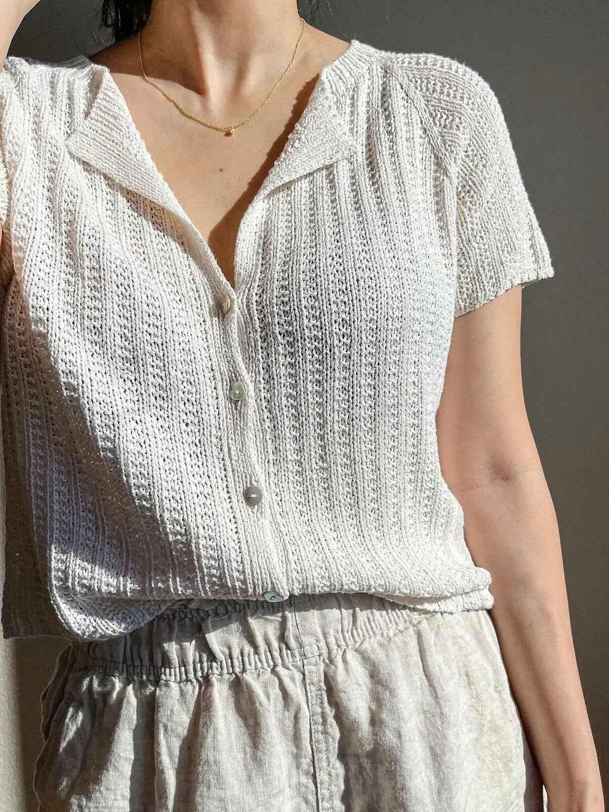 A person wearing a white, short-sleeved, button-up knit cardigan with a textured vertical pattern. The top features a small collar and is tucked into light-colored, high-waisted linen pants. The person is also wearing a delicate gold necklace.
