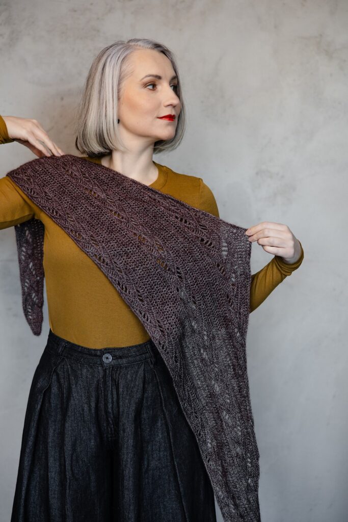 A woman with gray hair and red lipstick wears a mustard sweater and dark skirt, holding a purple-gray lace shawl. The setting has a neutral gray background.