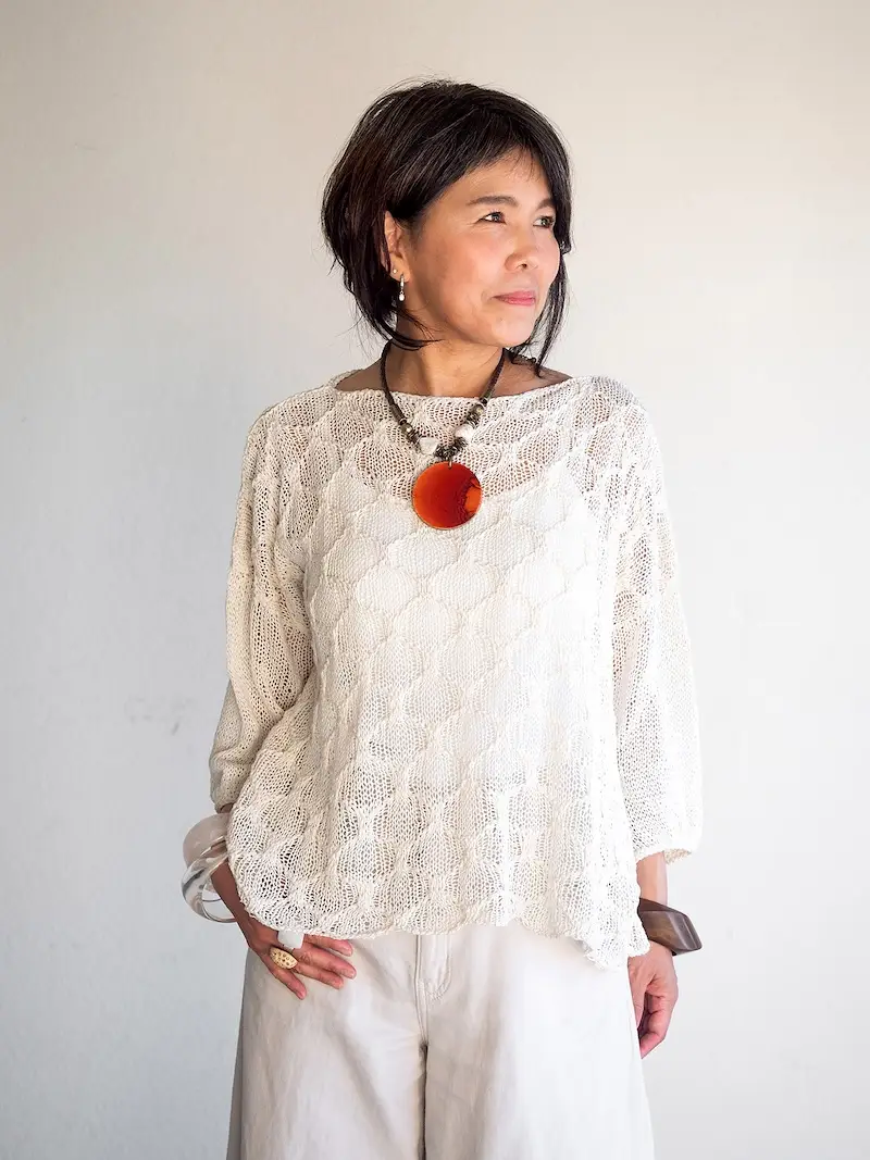 A woman wearing a white, loosely-knitted lace top with long sleeves, paired with a large circular orange pendant necklace. The top features an intricate, open-work pattern. 