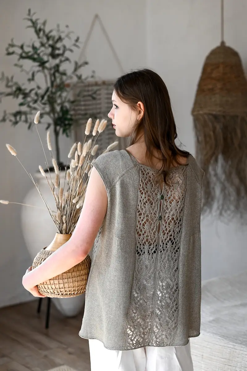 A woman facing away from us, wearing a sleeveless gray linen top with a buttoned lace panel running down the center of the back. She is standing in a minimalist, neutral-toned room, holding a small woven basket filled with dried wheat stalks. 