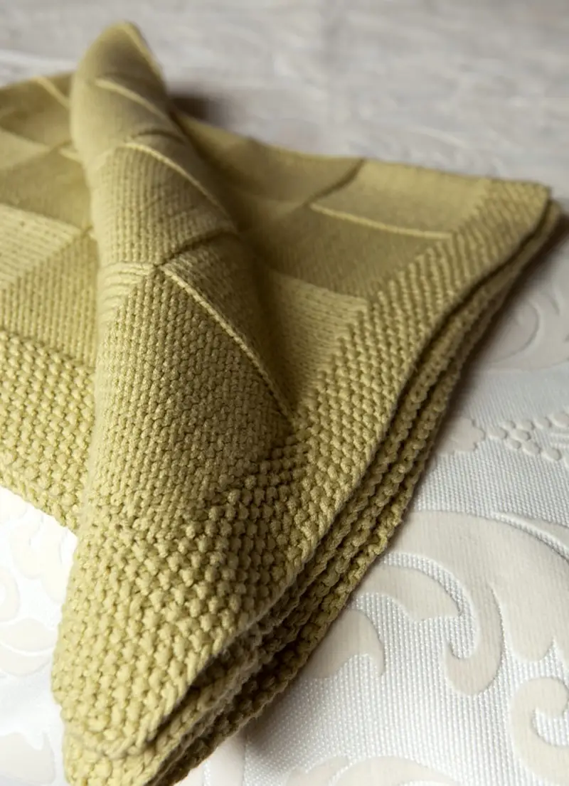 A folded yellow knitted baby blanket featuring a geometric pattern of textured squares. The blanket is finished with a wide seed stitch border and rests on a white patterned surface.