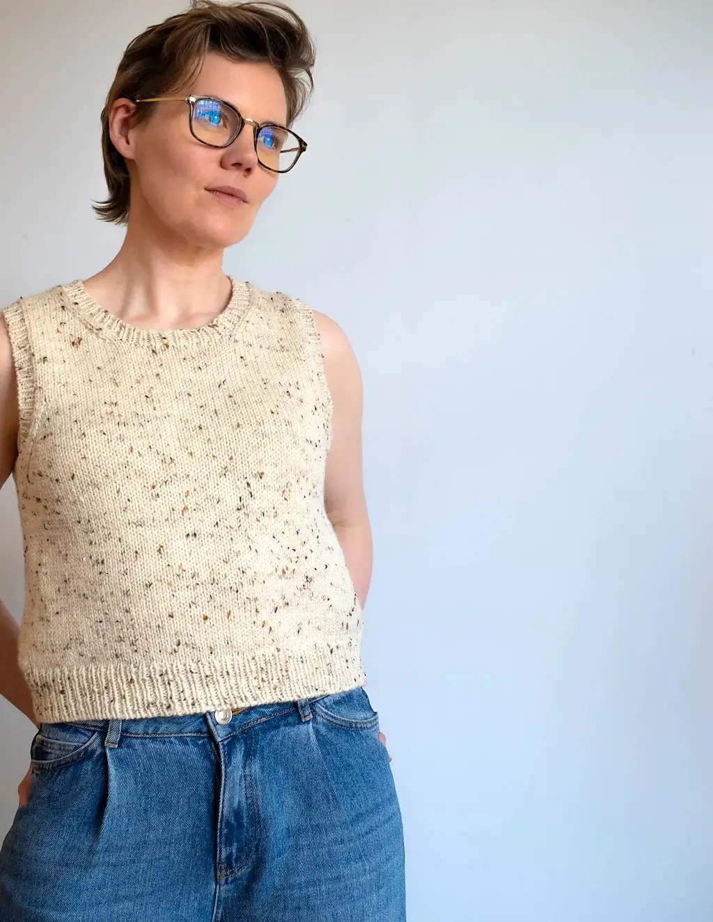 Ysolda Teague, a person with short brown hair and glasses looks off-camera, wearing a sleeveless, cream-colored knitted vest with dark flecks and light-wash blue jeans against a plain, light grey background.
