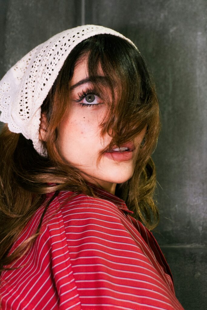 Person wearing a white lace headscarf and a red and white striped shirt, posing against a dark textured background.