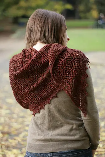 a woman with her back turned to the camera wears a large triangle lace shawl draped aound her shoulders knit in red-brown yarn. The lace design has a repeating diamond pattern.