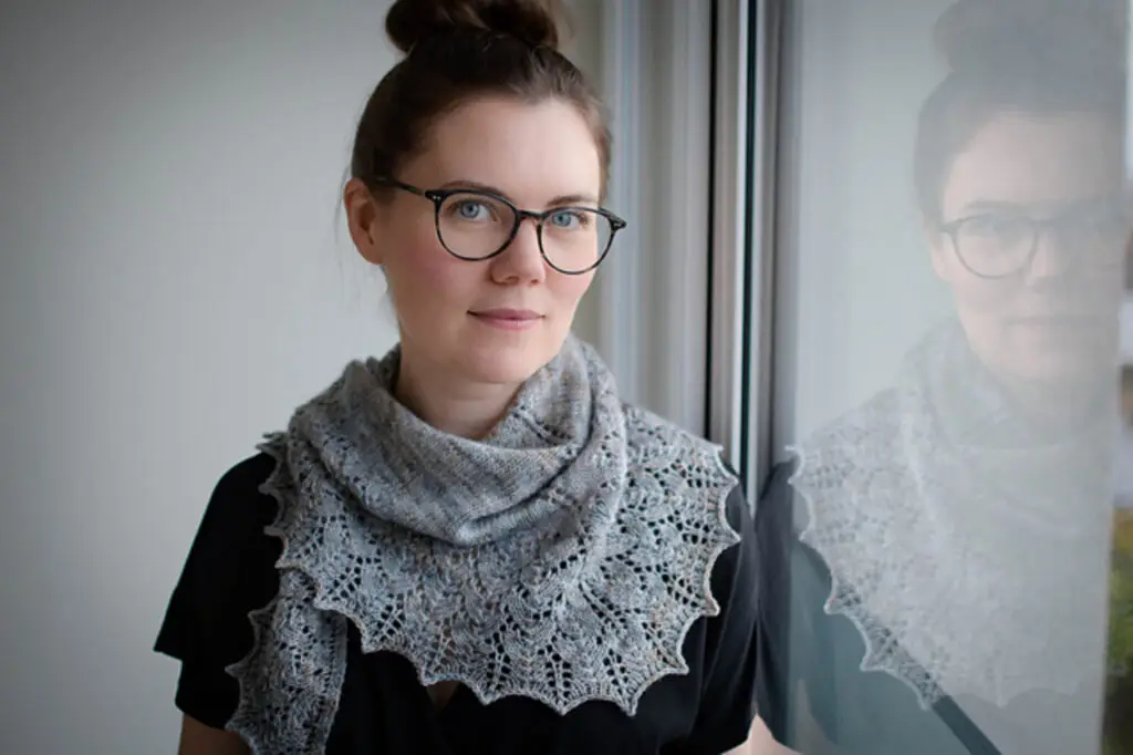 A woman in glasses with her hair in a bun faces the camera while leaning on a window. She wears a knit lace shawl made from a striking grey yarn wound around her neck.
