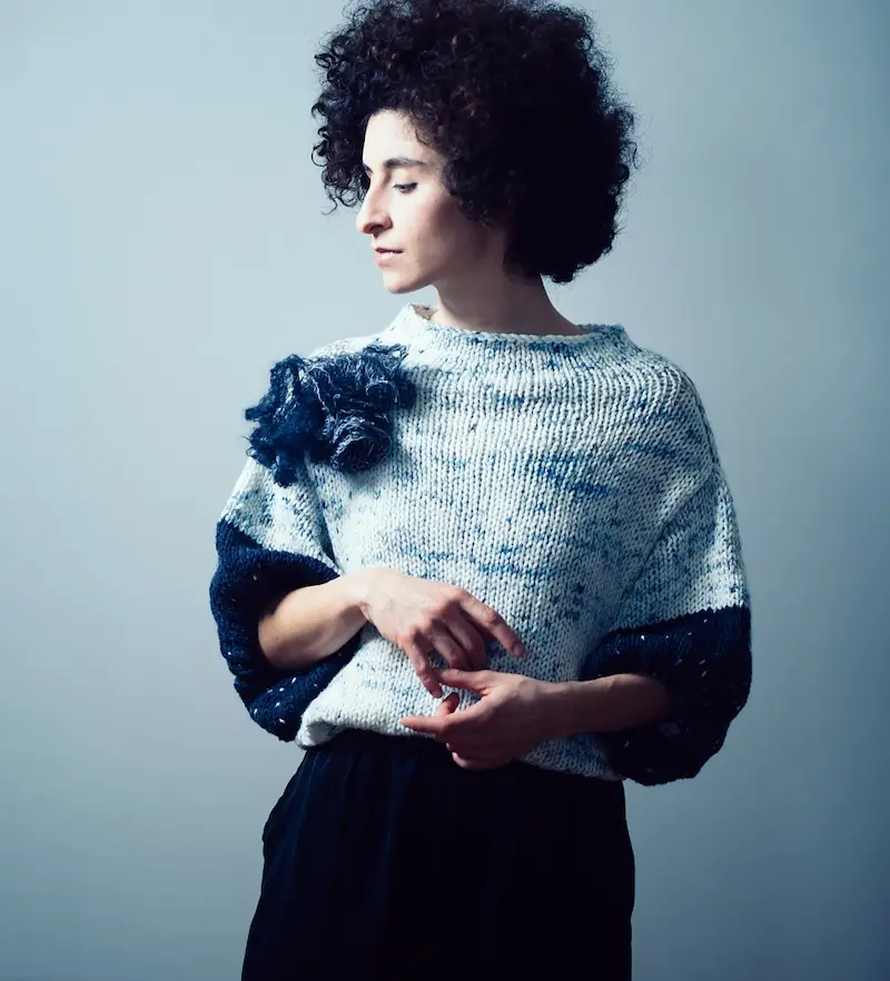 A woman with voluminous, dark, curly hair wearing a chunky hand-knit sweater in a pale yarn speckled with blue with contrasting dark blue 3/4 length sleeves. A prominent knitted floral embellishment is pinned to the right shoulder.