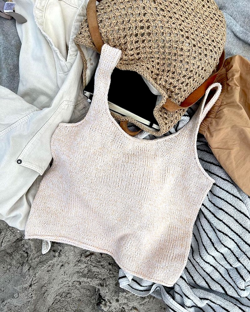 A cream-colored, hand-knitted tank top lies flat on a sandy beach, surrounded by beach essentials. The top features a wide, rounded neckline and thin straps, with a visible stockinette stitch pattern that creates a slightly textured, marled appearance in shades of off-white and pale beige.To the left of the top is a white button-down shirt, and to the right, a pair of tan shorts. Above the top sits a woven straw bag containing a black notebook. A striped white and navy beach towel is partially visible in the bottom right corner, all resting on the uneven, greyish-tan sand.