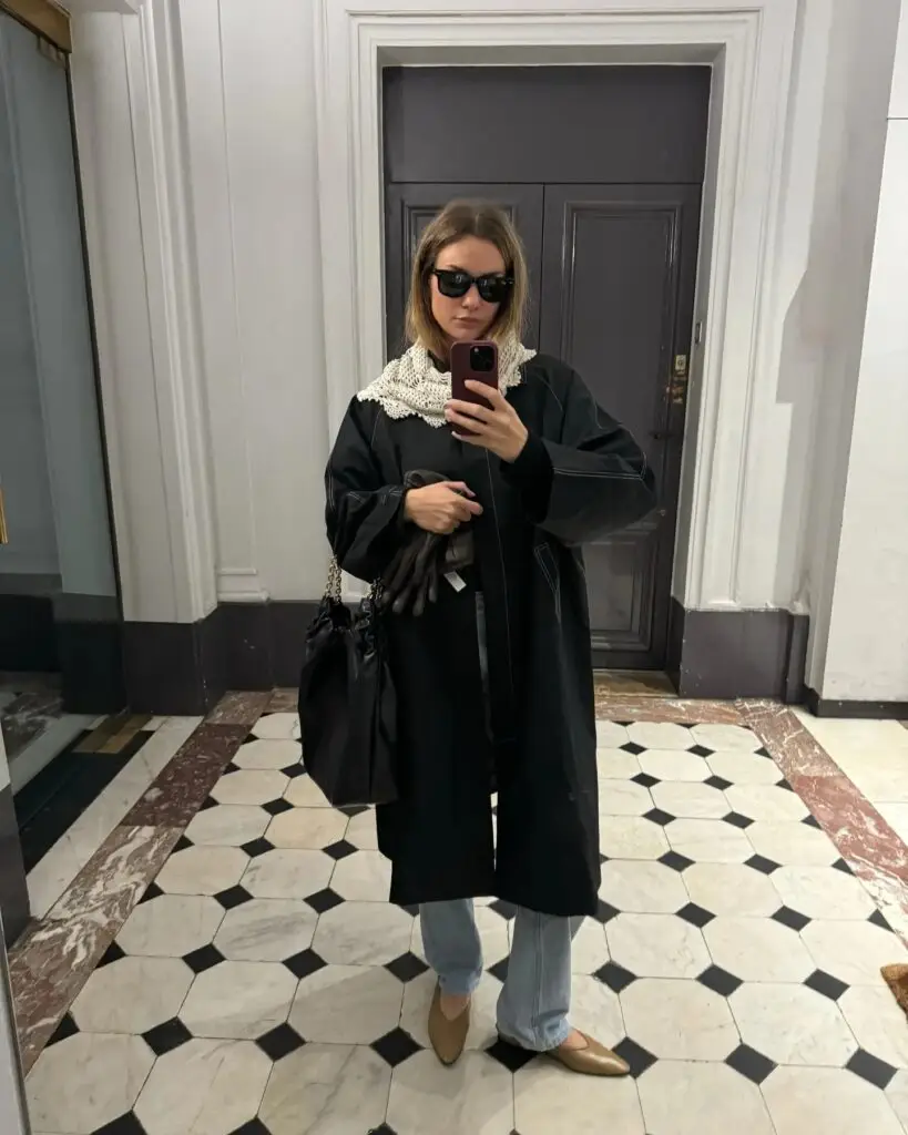 Person in a black coat, open stitched white scarf, blue jeans, and beige shoes taking a mirror selfie in a tiled entryway, holding gloves and a black bag.