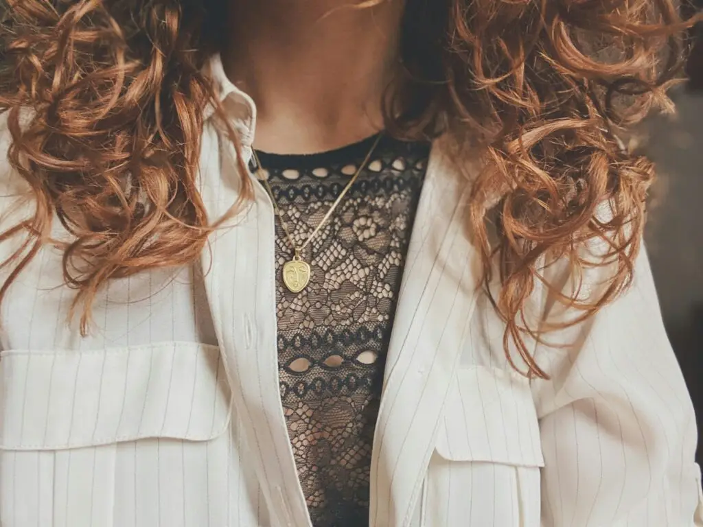 Close-up of a person with curly hair wearing a white pinstripe jacket over a patterned black lace top and a gold face pendant necklace.