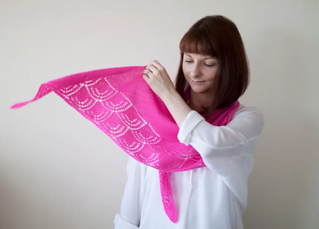 a woman with a bob and bangs wears a white buttondown shirt while actively throwing a magenta lace shawl around her shoulders. The lace design is scalloped along the edge
