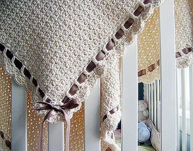 A cream colored textured crochet baby blanket is draped over the white rails of a crib. A thin brown satin ribbon is threaded through a row of decorative eyelet holes along the scalloped edge and tied into a delicate bow at the corner.