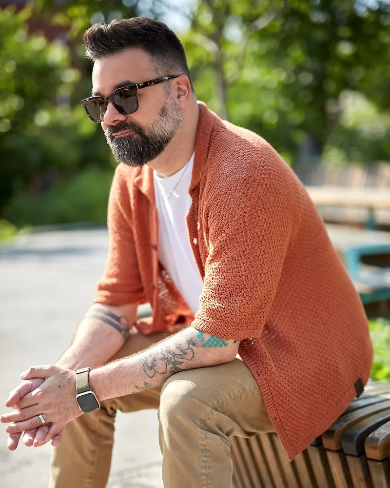 An adult man with a dark beard and sunglasses sits outdoors on a wooden bench. He is wearing an open, short-sleeved, rust-colored knit shirt over a white T-shirt and khaki pants.