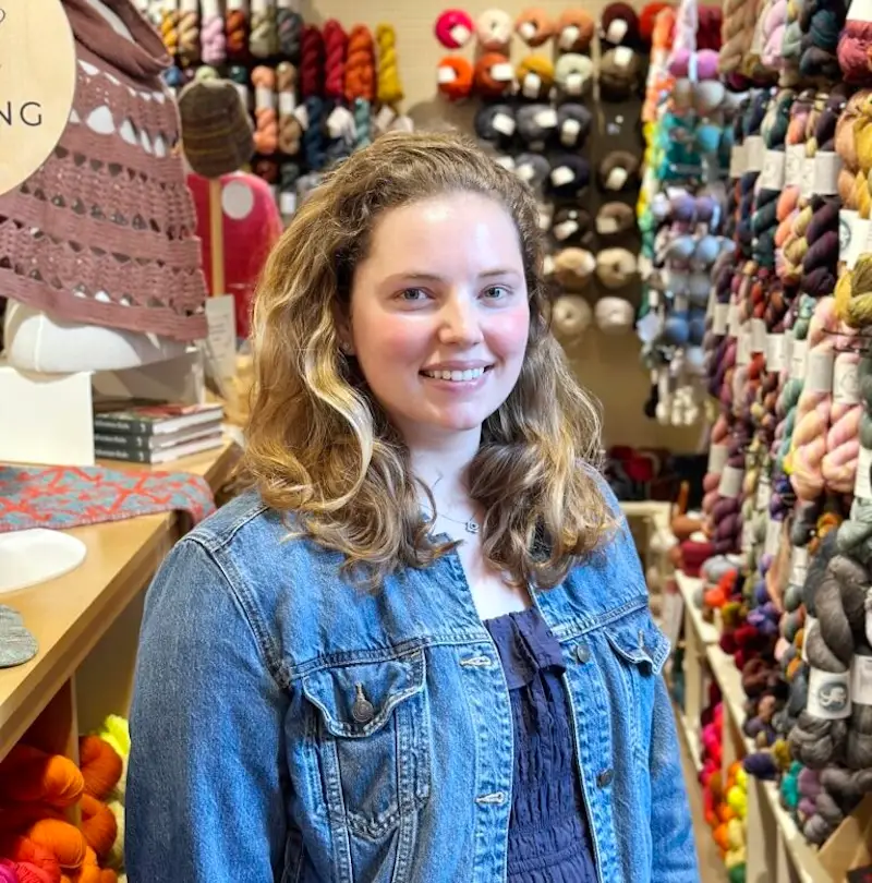 Jackson Roe, a young white woman with long dark blonde hair, stands in the aisle of fibre space surrounded by yarn.