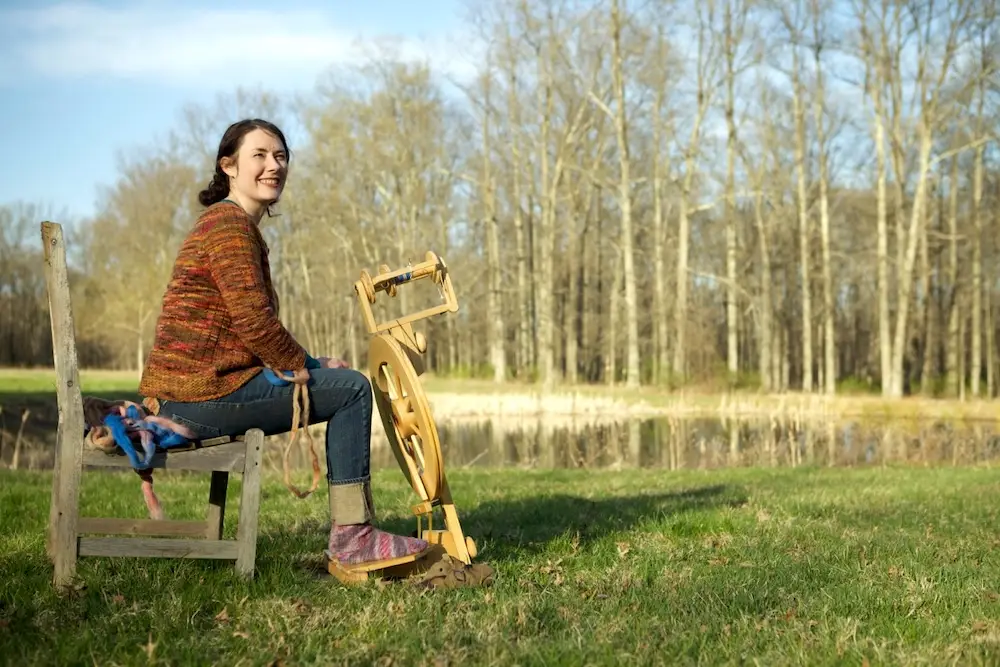Jill Draper, a smiling woman with light skin and long dark hair worn pulled back, sits outside with a wooden spinning wheel on the sunlit grassy shore of a pond surrounded by trees.