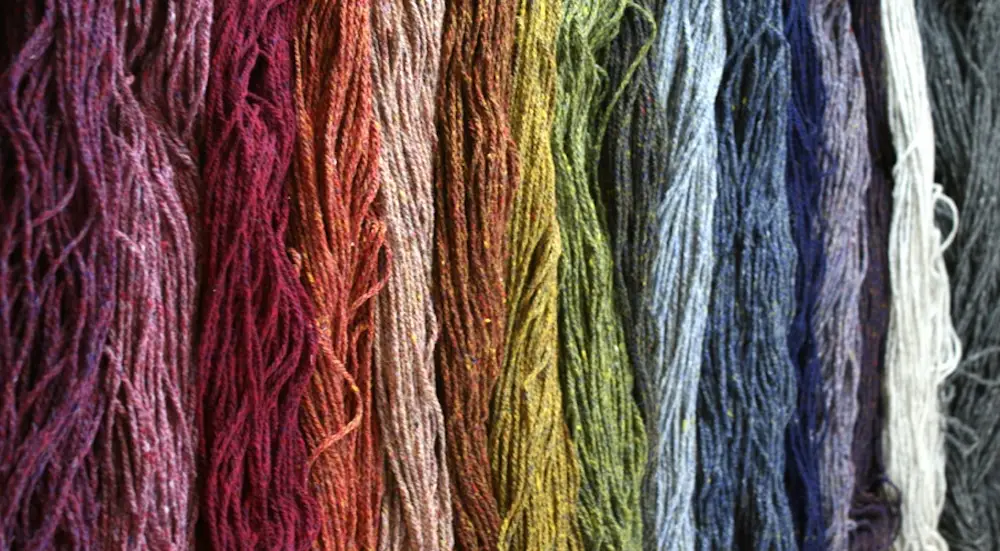 Loose skeins of heathered wool yarn hang side by side in colors ranging from warm pinks and reds on the left to cool blues, purples, and gray to the right.