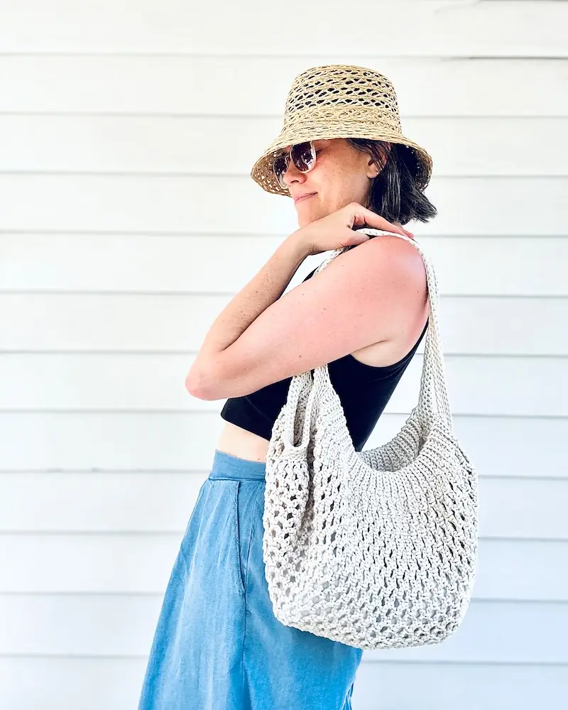 A person wearing a black tank top and straw hat carries a large white crocheted tote bag.