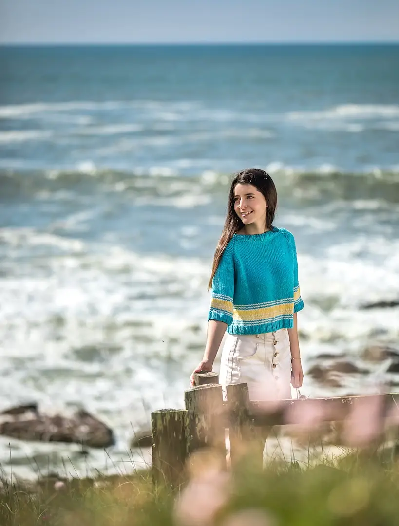 A young woman with long, dark hair stands outdoors near the ocean. She is wearing a teal, short-sleeved knitted sweater with two yellow horizontal stripes near the hem and a white, button-front skirt. She leans lightly against a rustic wooden post. The background features a vast, churning sea with white-capped waves under a clear sky, with blurred green grass and pink flowers in the foreground.