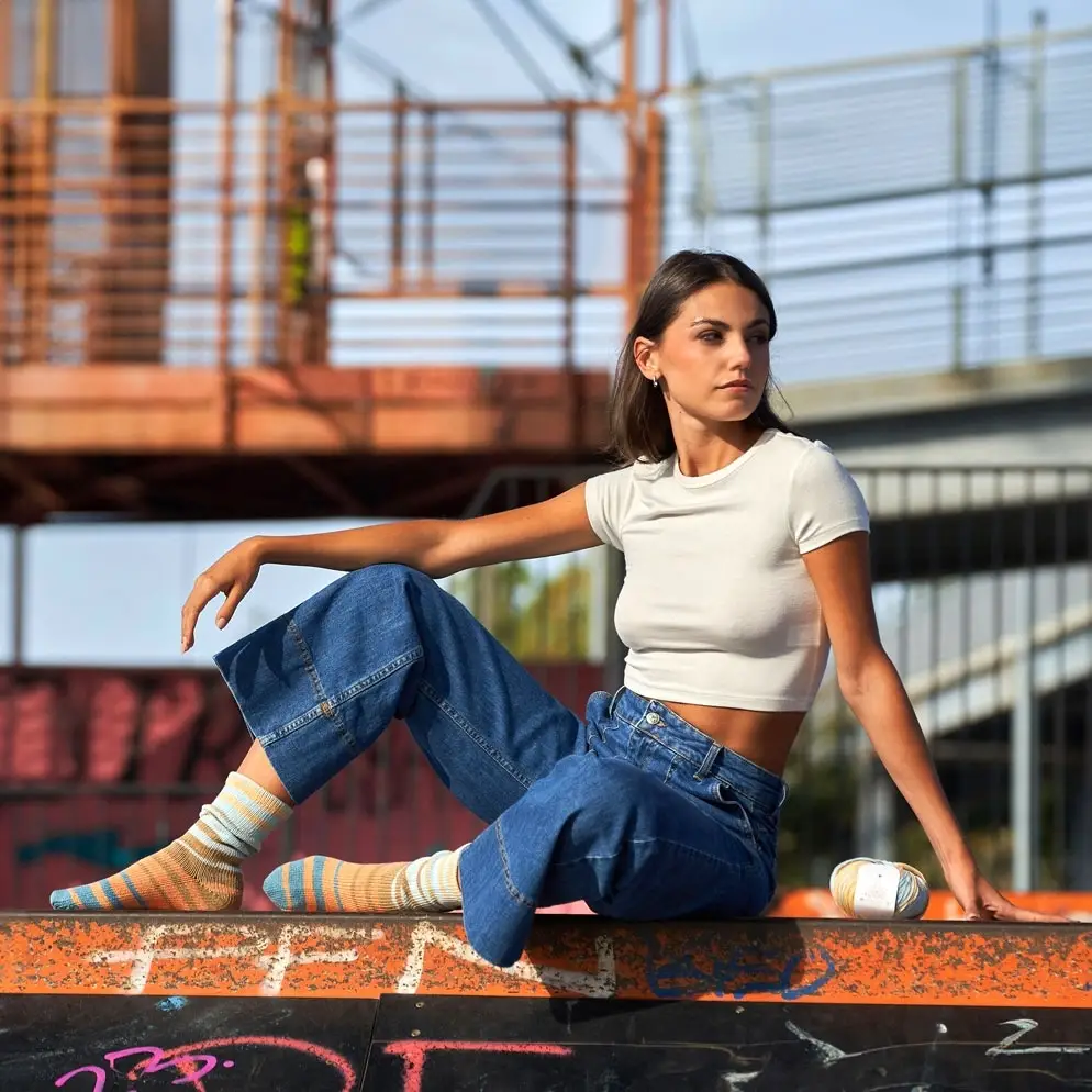 A woman sits outdoors on a ledge below an orange metal bridge wearing a white cropped t-shirt, wide-leg blue jeans, and colorful striped knit socks. Next to her on the ledge rests a skein of self-striping yarn in matching shades of blue, beige, and orange.