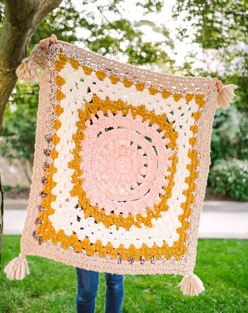 A person stands under a tree holding up a chunky crocheted granny square blanket in tan, gold, cream, and pink.