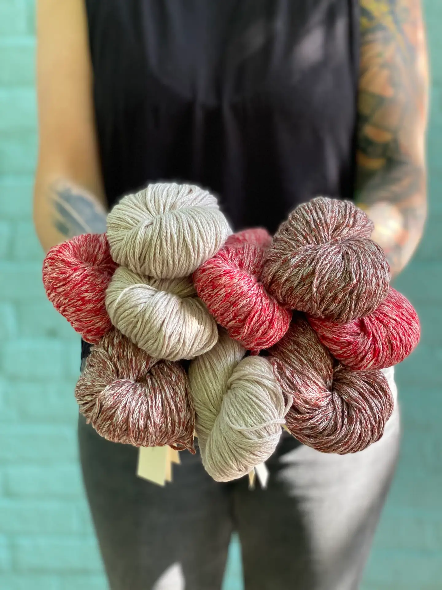 Person with tattoos holding multiple skeins of red, pink, beige, and brick Woolfolk Brise yarn against a turquoise brick background.