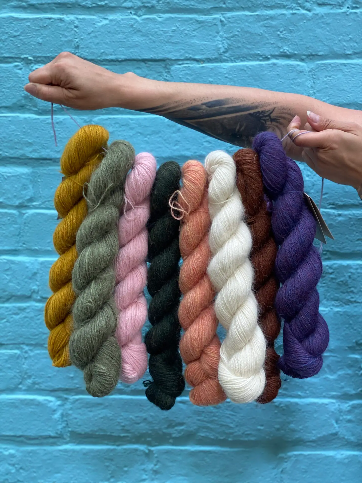 Person holding eight colorful skeins of Baa Ram Ewe Mowool in front of a blue brick wall, showcasing a range of earthy and vibrant tones.