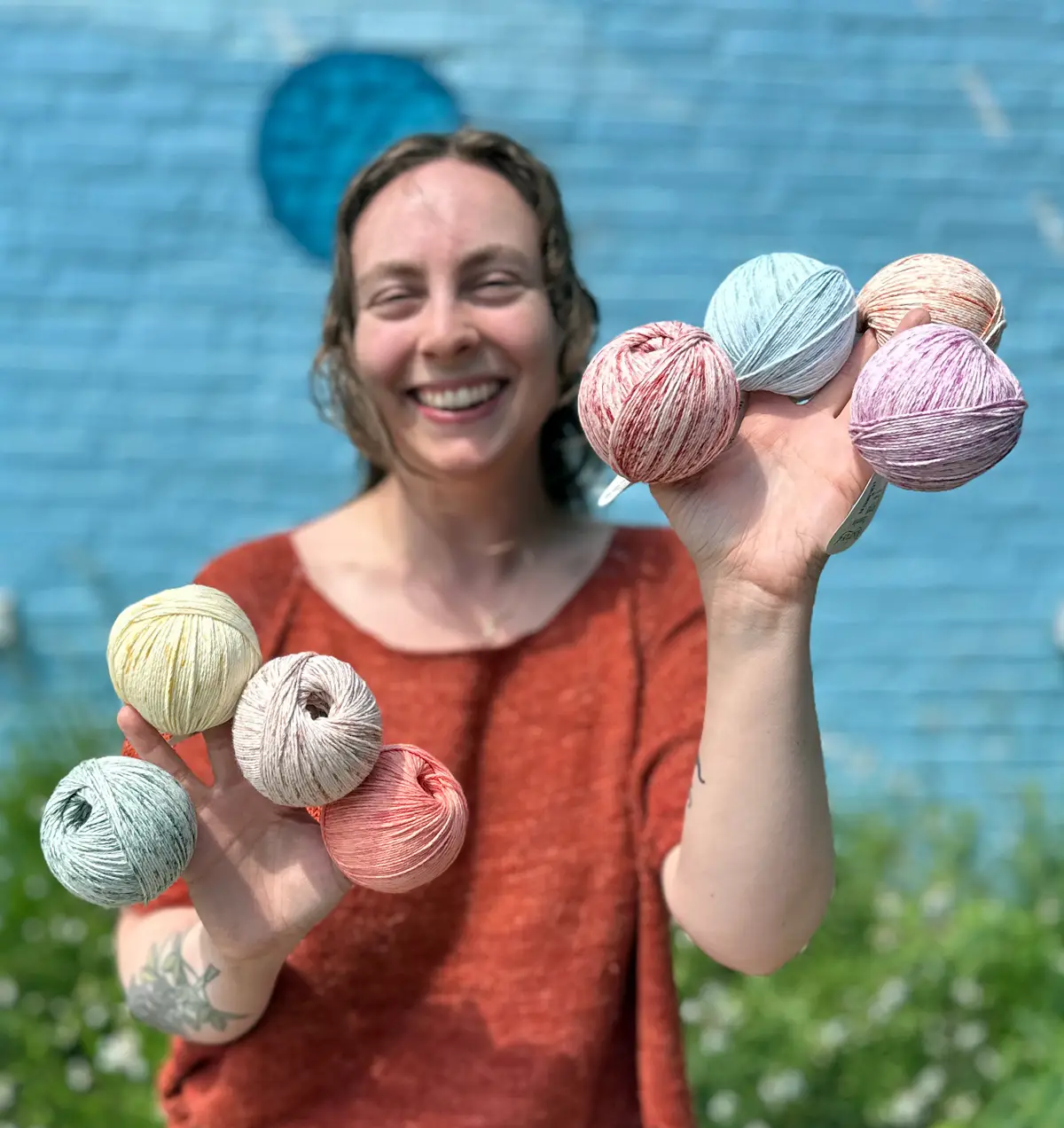 A woman holds speckled and colorful balls of Rosários 4 Odemira in both hands. She stands against a blue brick wall background with greenery below.