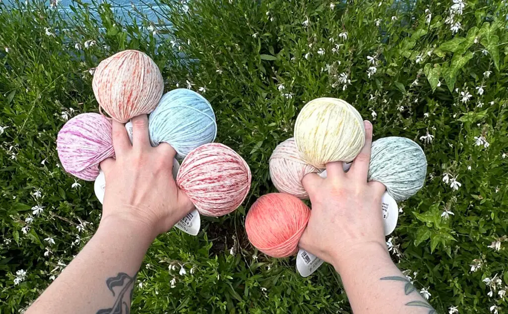A person's hands holds multiple speckled and colorful balls of Rosários 4 Odemira yarn, over green foliage with small white flowers.