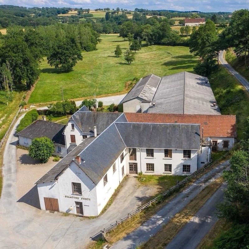 Aerial view of a the historical Fonty spinning mill building in France, with multiple buildings surrounded by green fields and trees under a partly cloudy sky.