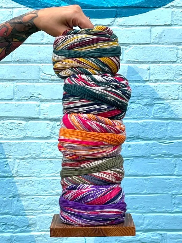 A person with tattoos holds the top of a wooden yarn ball holder stacked with big multicolored balls of sock yarn.