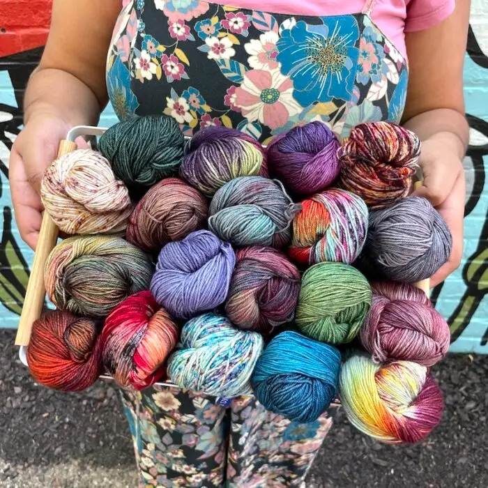 A person in colorful floral-print overalls holds a basket filled with multicolored skeins of Malabrigo Sock yarn.