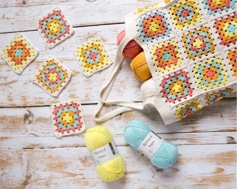 Skeins of Rosários 4 Regata yarn spill out of a colorful granny square tote bag.