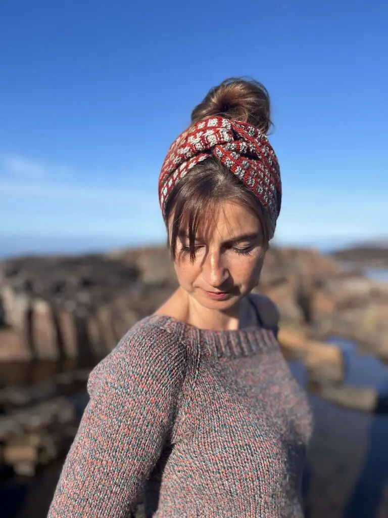 Pattern designer Brienne Moody models a patterned headband while outdoors near rocky water under a clear blue sky.