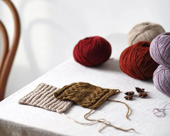 A linen-covered table holds balls and swatches of De Rerum Natura yarns in deep red, warm brown, and heathered purple.