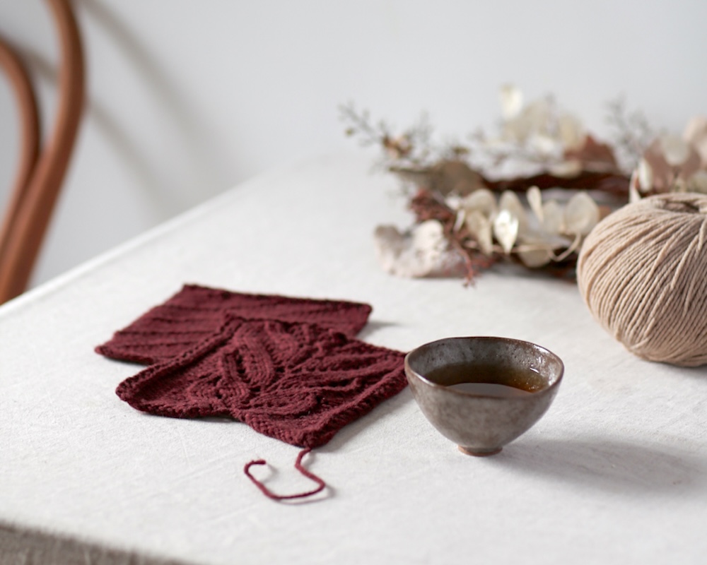 Balls and swatches of De Rerum Natura Ulysse yarn in warm beige and deep red on a linen-covered table.