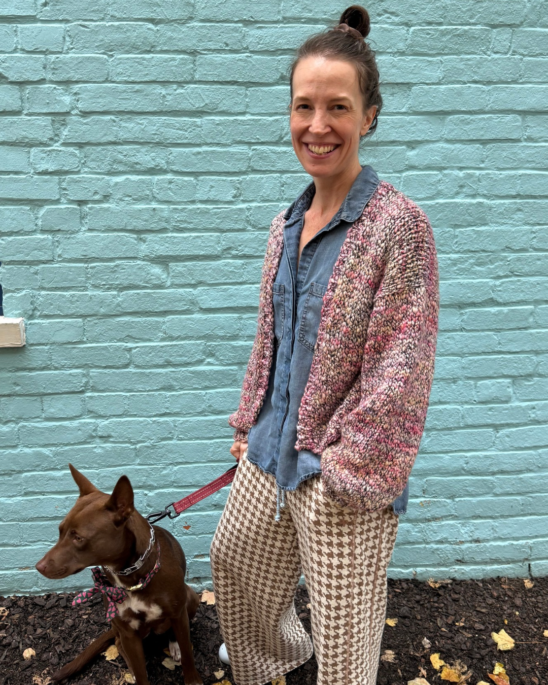 Danielle wears the Oli Cardigan in a mostly light pink variegated shade of Malabrigo Caracol. She stands in front of fibre space's blue wall, along with her dog Mocha.